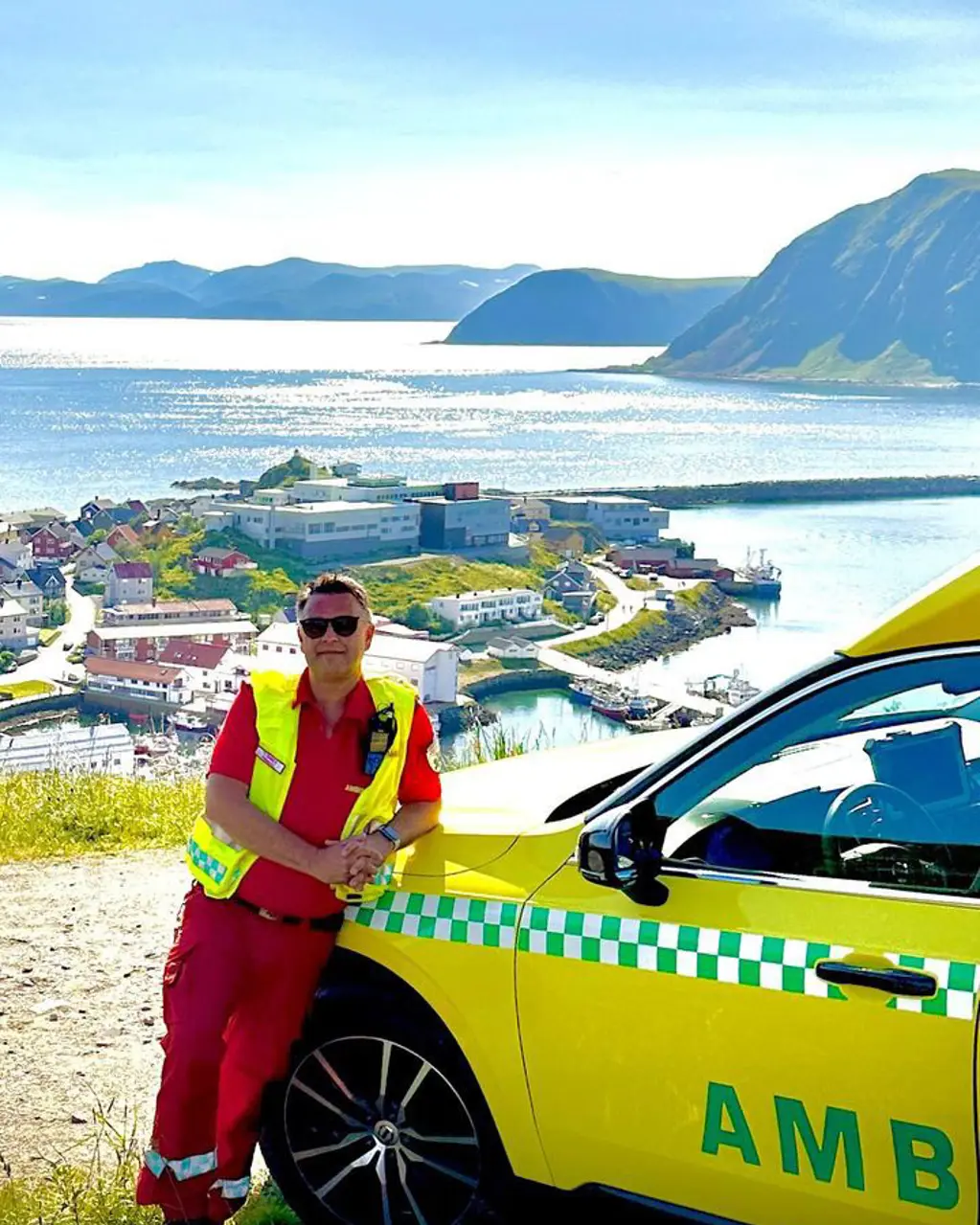 En mann i redningsvest som står ved siden av en gul bil på en strand med en by og