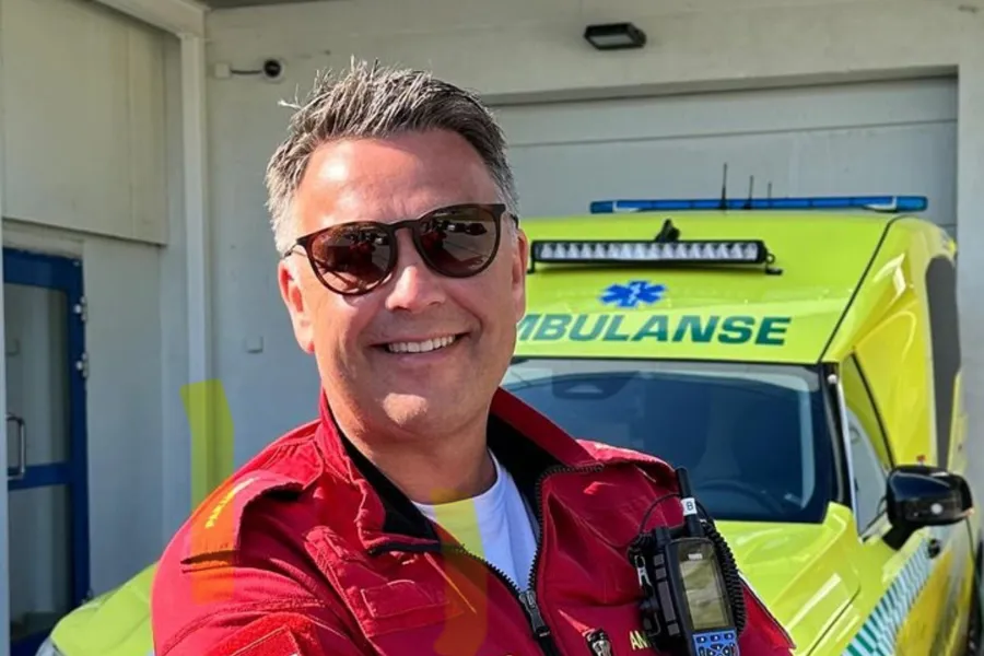 En mann iført solbriller og en rød jakke med en gul bil bak seg